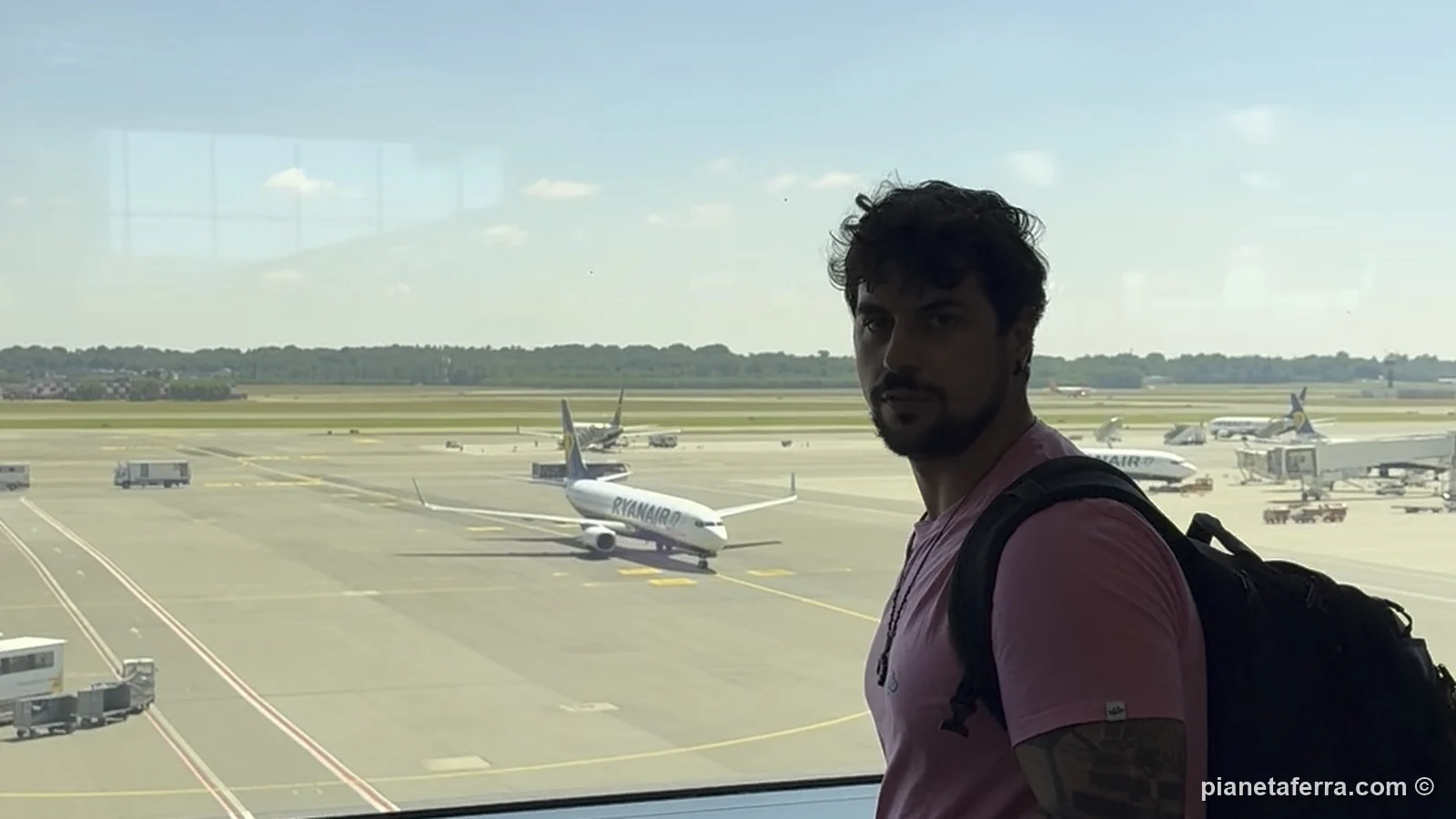 Luca con lo zaino davanti alla vetrata dell'aeroporto di Malpensa, con gli aerei sulla pista, nell'estate del 2025