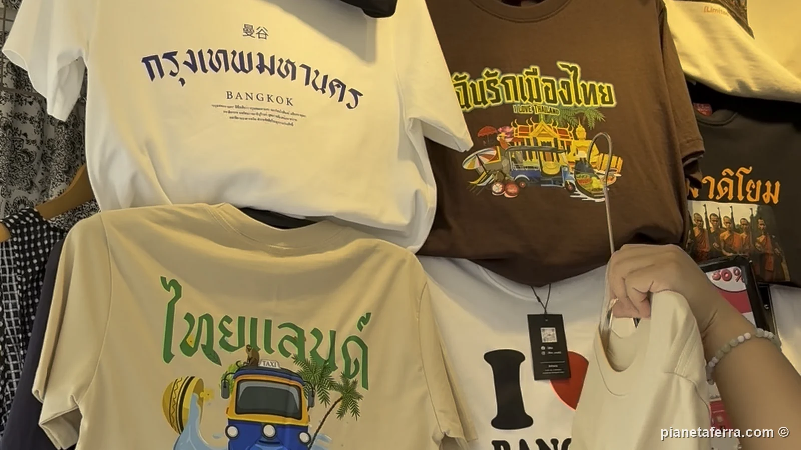 Magliette con scritte in thailandese e disegni di tuk tuk appese a un banco del mercato di Chatuchak