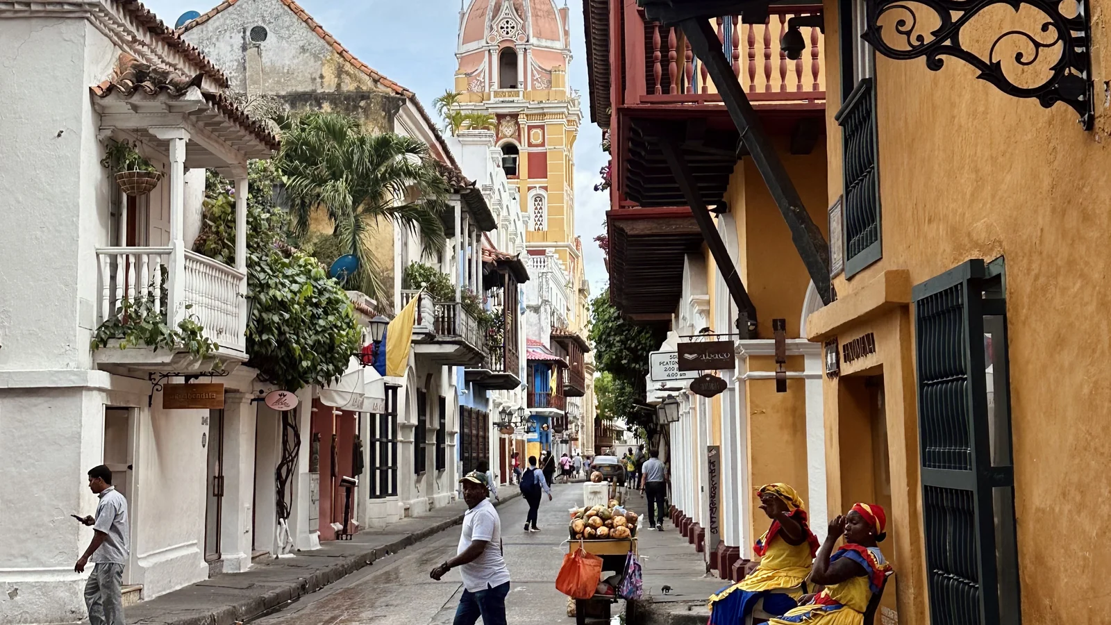 Vicolo coloniale di Cartagena con balconi in legno, campanile della cattedrale e palenqueras con la bandiera colombiana