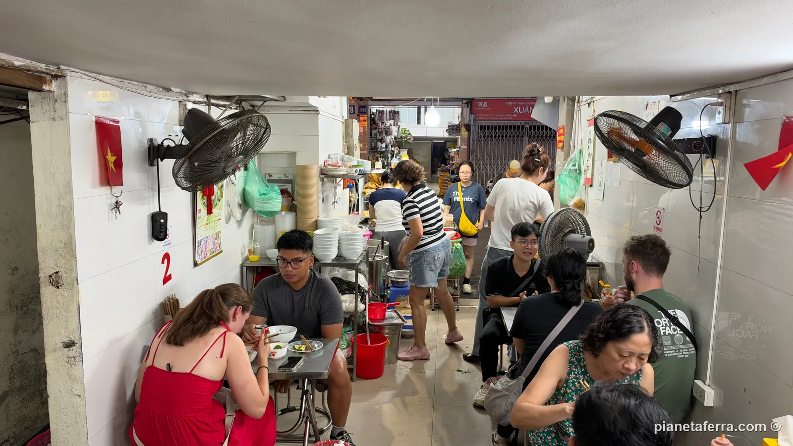 La sala al piano terra di Phở Hà ad Hanoi, un corridoio stretto con i tavolini di metallo, i ventilatori alle pareti e la cucina in fondo