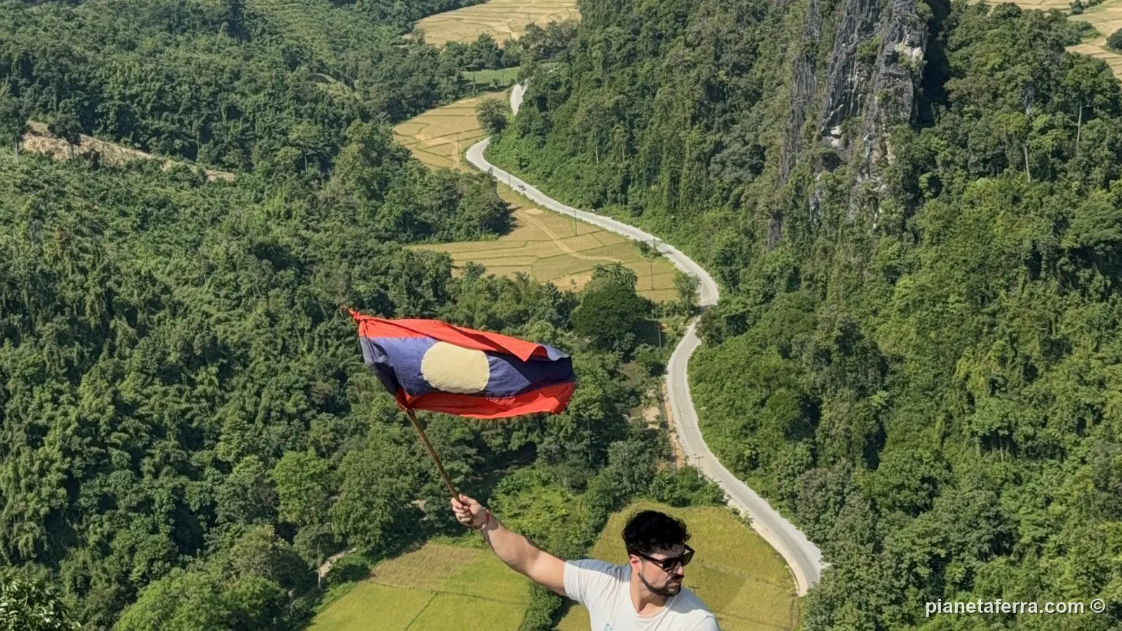 Luca sventola la bandiera del Laos dal Nam Xay Viewpoint, con la strada e le risaie sotto